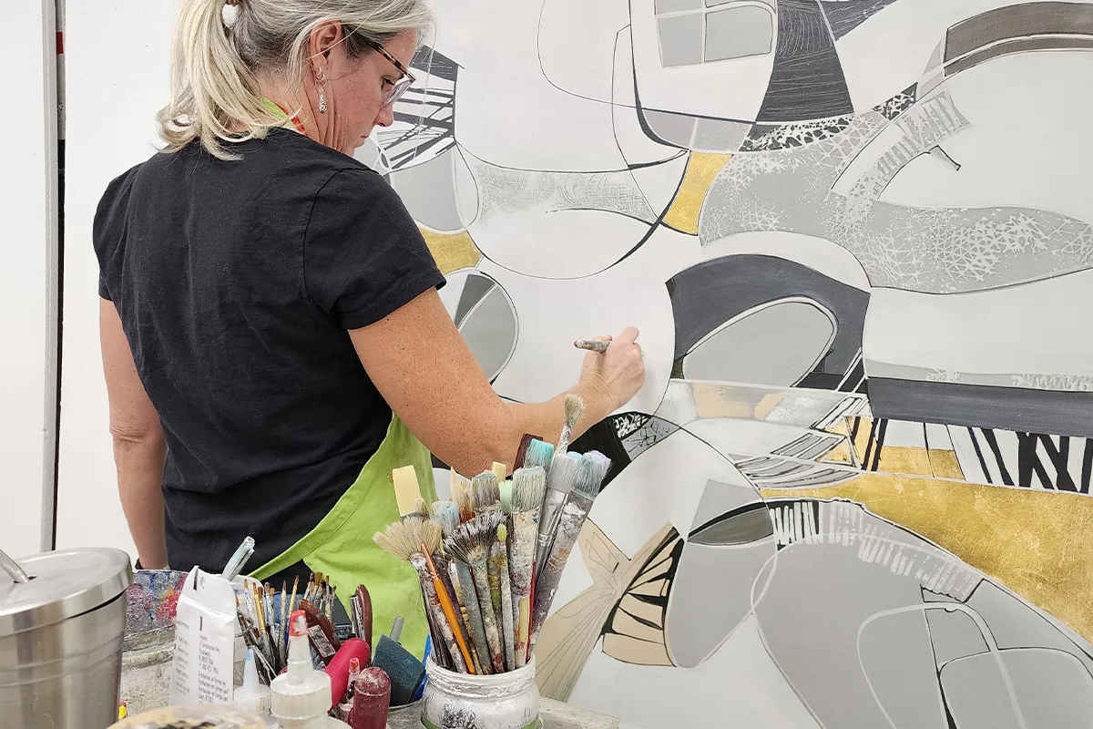 Female artist embellishing a contemporary abstract print with gold and neutral tones, adding texture and dimension using fine brushes in a professional art studio.