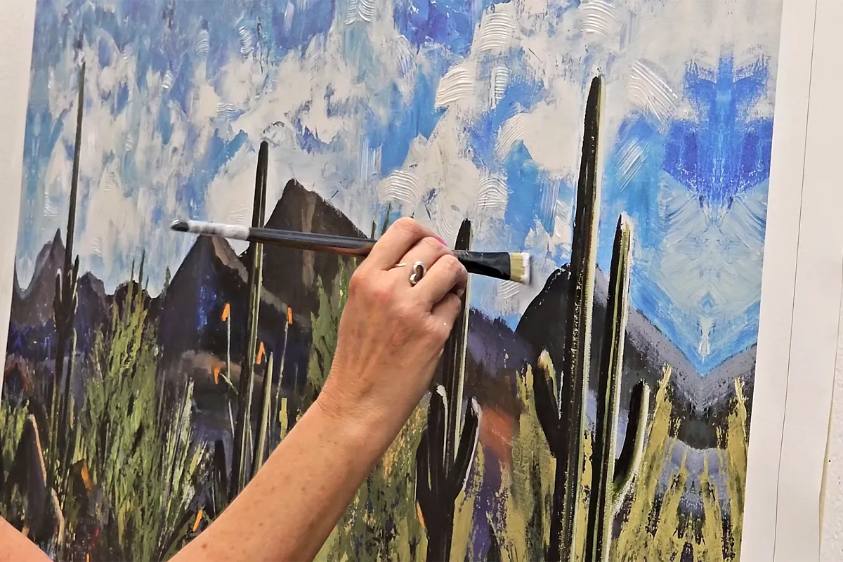 Close-up of artist embellishing a printed desert landscape featuring saguaro cacti and mountains under a textured blue sky, enhancing depth and brushwork in a fine art studio setting.